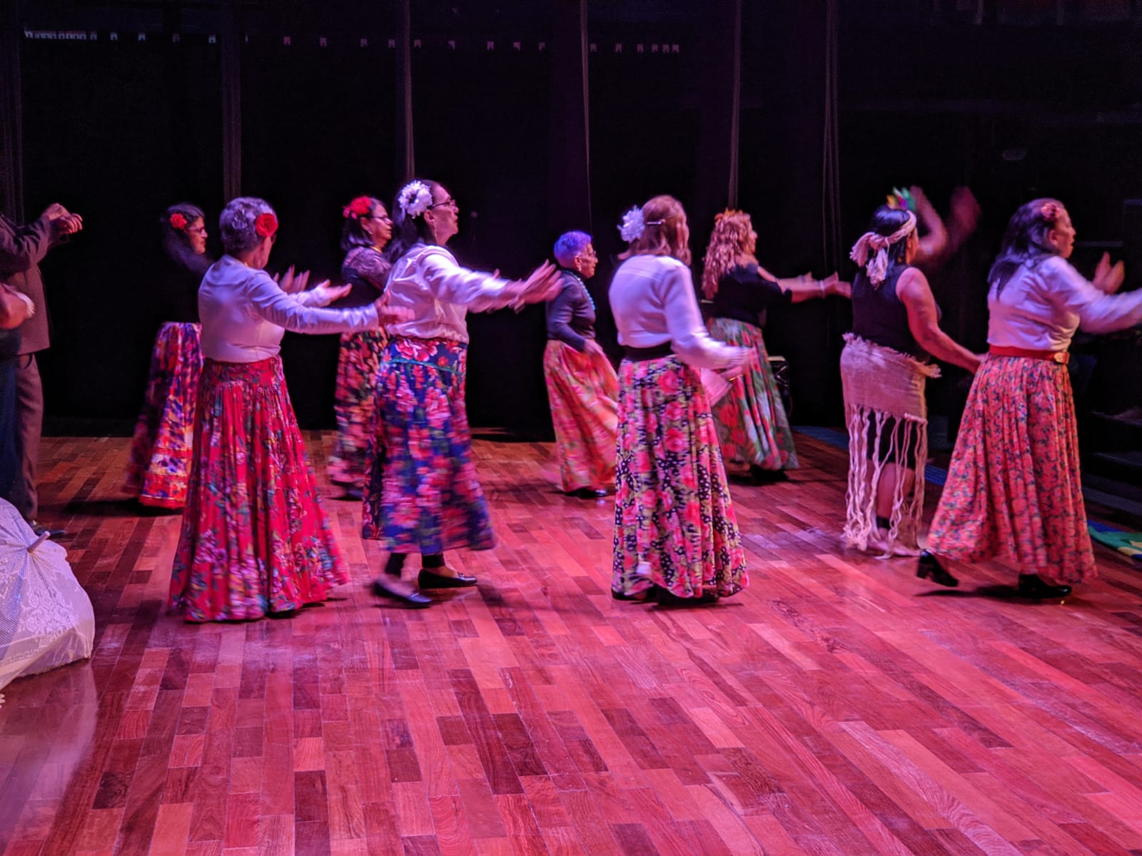 Na imagem, um grupo de idosas realiza uma dança em cima do palco, com uma luz rosa forte em cima delas. Elas vestem saias longas e floridas, e flores nas cabeças.